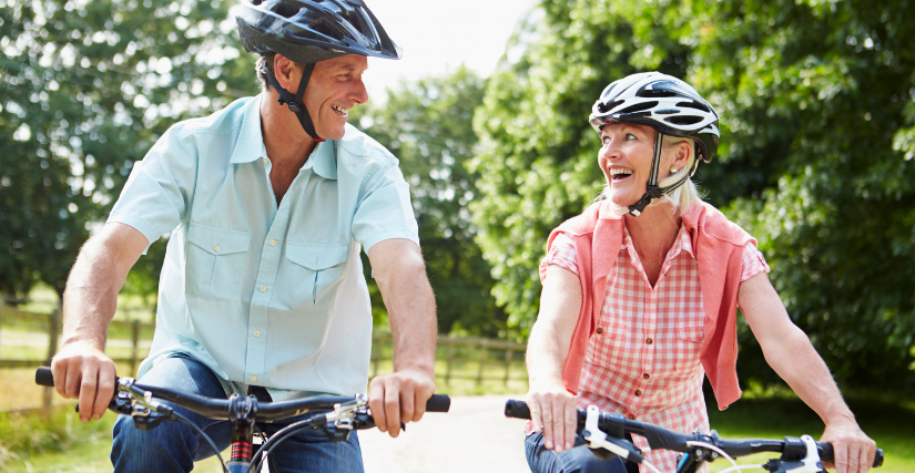 A smiling, active mature couple riding bicycles side by side along a dirt path on a sunny day. Both are wearing black and white bicycle helmets. The man on the left wears a light blue short-sleeved button-down shirt, and the woman on the right wears a pink plaid shirt with a coral sweater draped over her shoulders. Lush green trees and a wooden fence line the background.