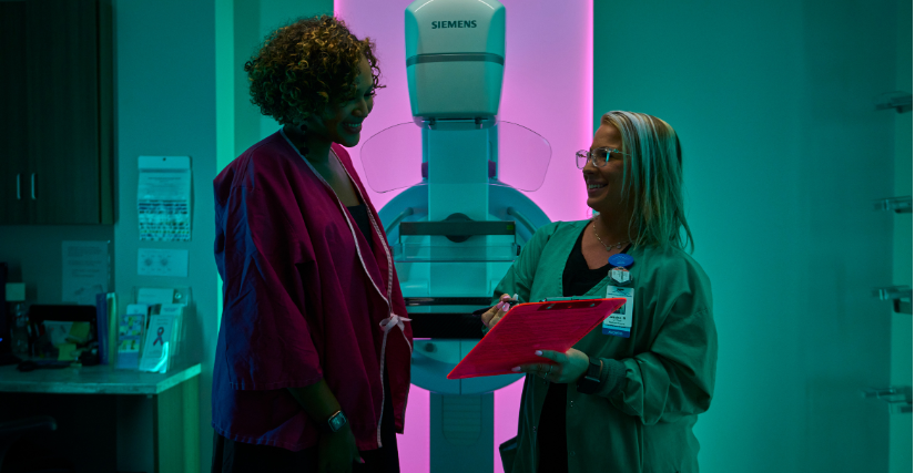 A healthcare provider wearing glasses, green scrubs, and an ID badge smiles while holding a pink clipboard and taking notes. She stands in a brightly lit medical room beside a female patient wearing a pink patient gown. In the background between them is a Siemens mammography machine illuminated with pink and teal accent lighting.