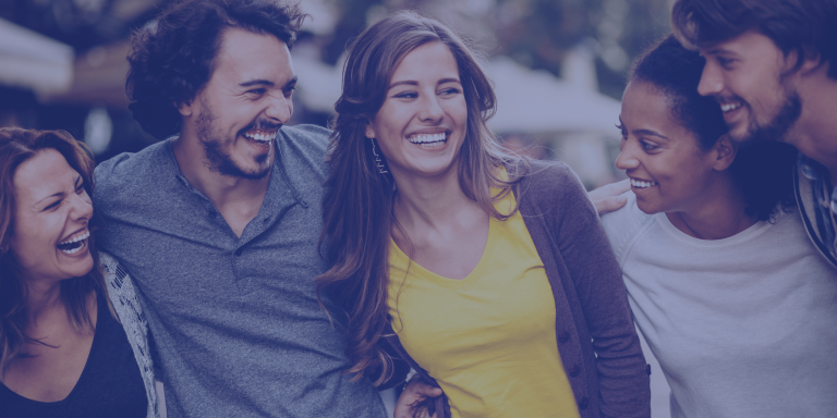 Group of five diverse young adult friends walking together outdoors, laughing with their arms around each other.