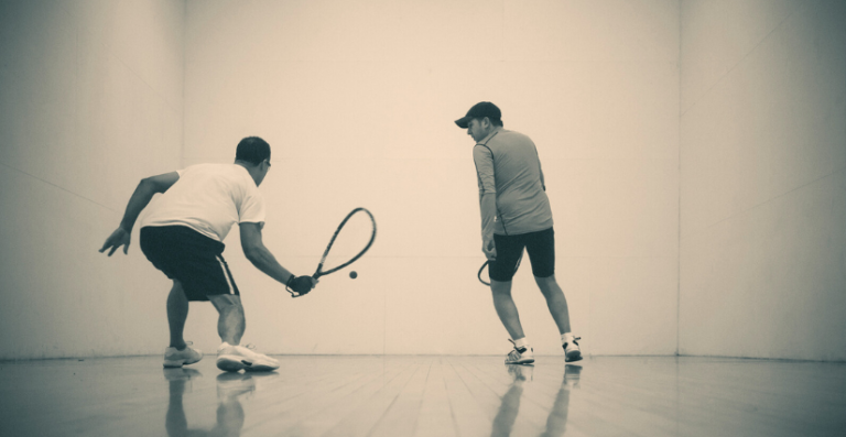 Two men playing racquetball inside an indoor court, seen from behind.