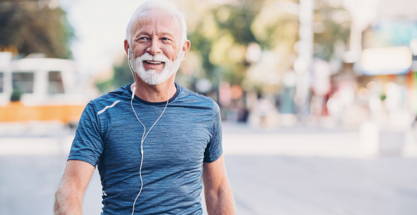 Smiling senior man with a white beard wearing a blue athletic shirt and wired earphones, walking outdoors on a sunny day.