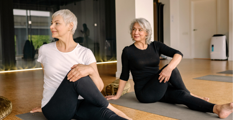 Two senior women practicing a seated spinal twist yoga pose on mats in a bright studio.