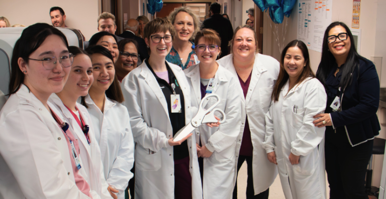 A diverse group of about ten healthcare workers smiling together in a brightly lit facility corridor. Most team members wear white lab coats over scrubs or professional attire with ID badges, standing close together for a group photo celebration.