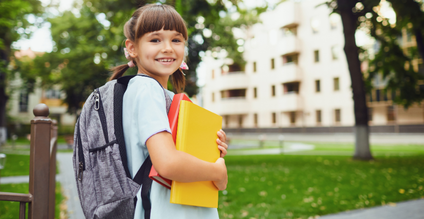 A young girl with pigtails wearing a grey backpack and holding bright yellow and red books against her chest, smiling at the camera outdoors in a park or school setting.